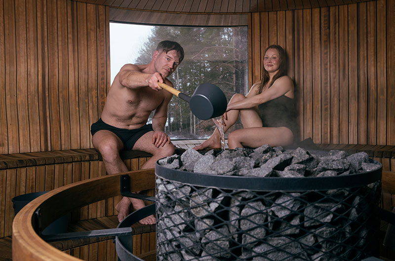 Muž leje vodu naberačkou na rozpálené saunové kamene, žena sedí neďaleko; parná, okrúhla drevená sauna s oknom.